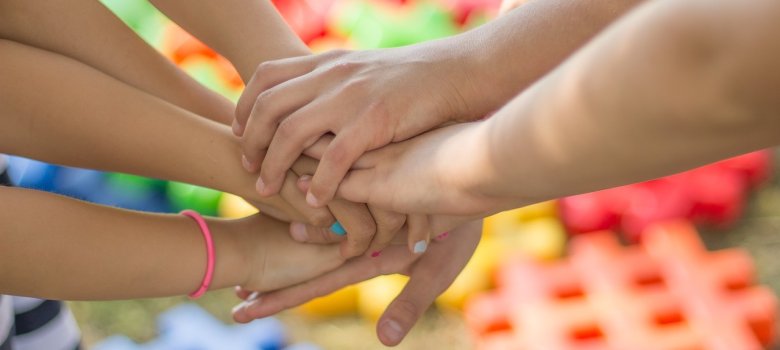 Mehrere Arme und Hände von Kindern liegen in der Bildmitte als Zeichen von Gemeinschaft übereinander gestapelt. Im unscharfen Hintergrund sind bunte Spielsteine aus Kunststoff auf einer Rasenfläche zu erkennen