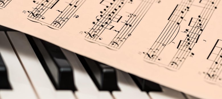 Draufsicht auf eine Klaviertastatur im Anschnitt; darauf liegt schräg ein Notenblatt mit komplexer Klaviernotation. Der Fokus liegt auf den Notenlinien im Vordergrund, während die Tasten leicht unscharf verlaufen
