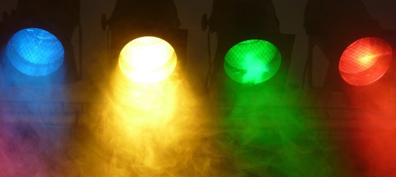 Vier Bühnenscheinwerfer sind nebeneinander aufgereiht und leuchten in den Farben Blau, Gelb, Grün und Rot. Das Licht tritt in dichten Strahlen nach unten aus und wird durch künstlichen Nebel sichtbar gemacht, der die Farben weich ineinanderfließen lässt
