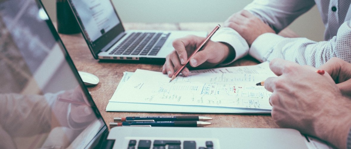 Nahaufnahme einer Arbeitssituation: Im Vordergrund steht ein offener Laptop, dahinter schreibt eine Person mit einem Stift auf ein Dokument, während eine zweite Person daneben sitzt. Ein weiterer Laptop ist im Hintergrund sichtbar