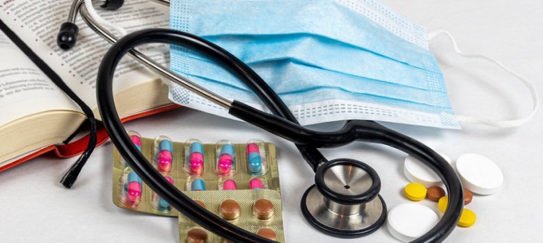 Nahaufnahme von medizinischem Zubehör: Ein schwarzes Stethoskop liegt auf einer OP-Maske und Blisterpackungen mit pink-blauen Kapseln; daneben ein offenes Buch mit engem Text