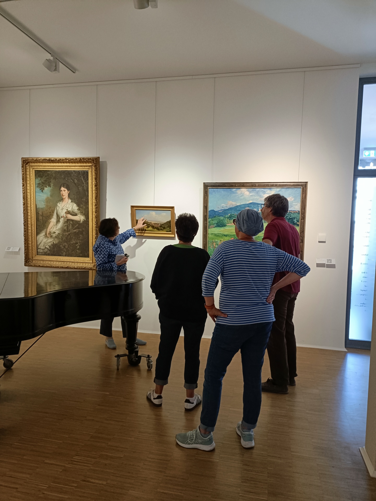 Führung Führung durch ein Kunstmuseum oder eine Galerie auf hellem Parkettboden. Eine Museumsführerin steht links vor einem großen, schwarzen Flügel und zeigt auf ein kleineres, gerahmtes Landschaftsgemälde an der weißen Wand. Eine dreiköpfige Besuchergruppe steht im Vordergrund von hinten betrachtet und hört aufmerksam zu. Links und rechts an der Wand hängen zwei weitere große, klassisch gerahmte Ölgemälde – ein Frauenporträt und eine Landschaft.