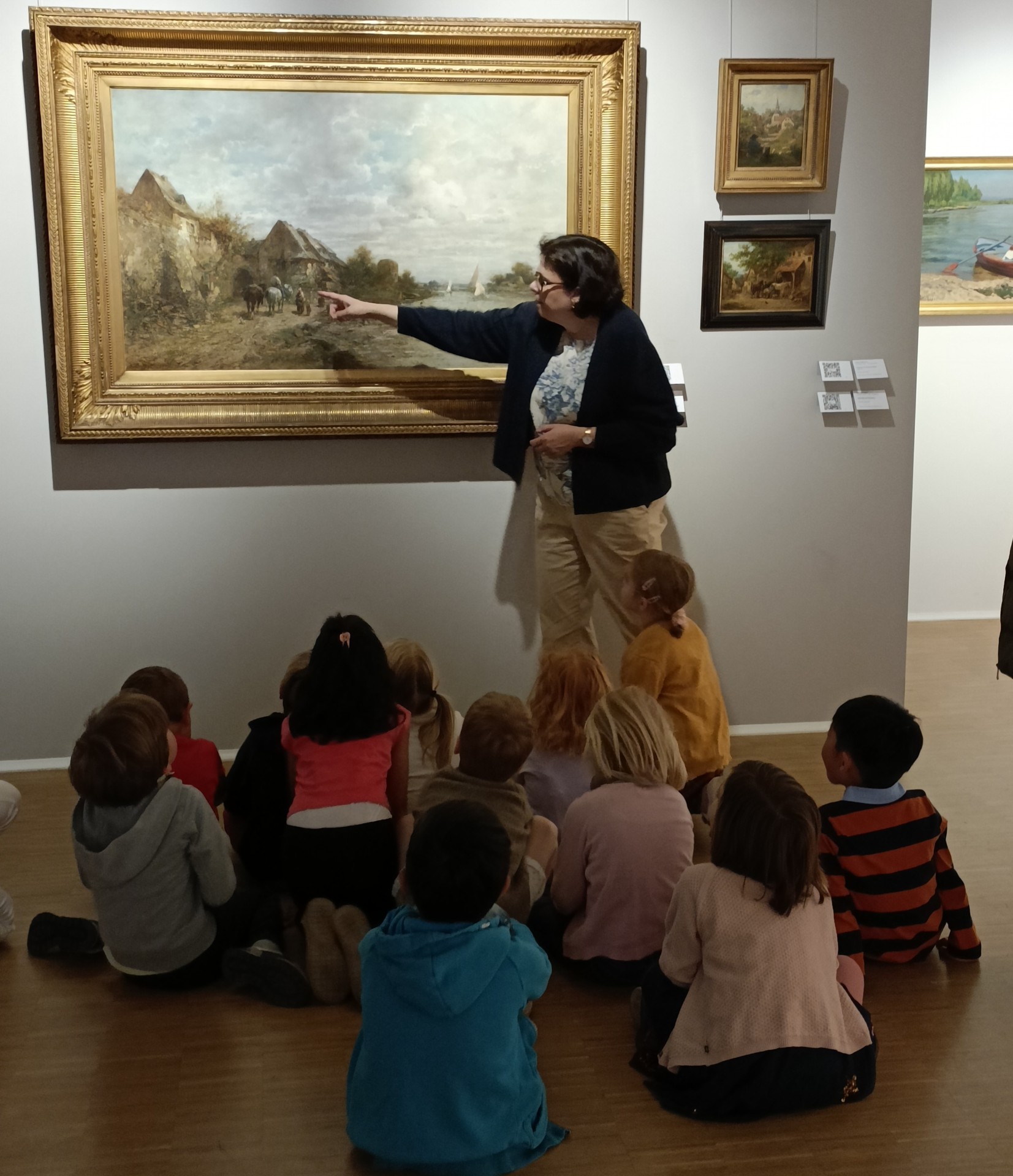 Eine Museumsführerin zeigt einer Gruppe von Kindern, die auf dem Boden sitzen, ein großes Landschaftsgemälde in einem goldenen Rahmen. Die Frau deutet auf ein Detail im Bild, während die Kinder aufmerksam zu ihr und dem Kunstwerk aufblicken.