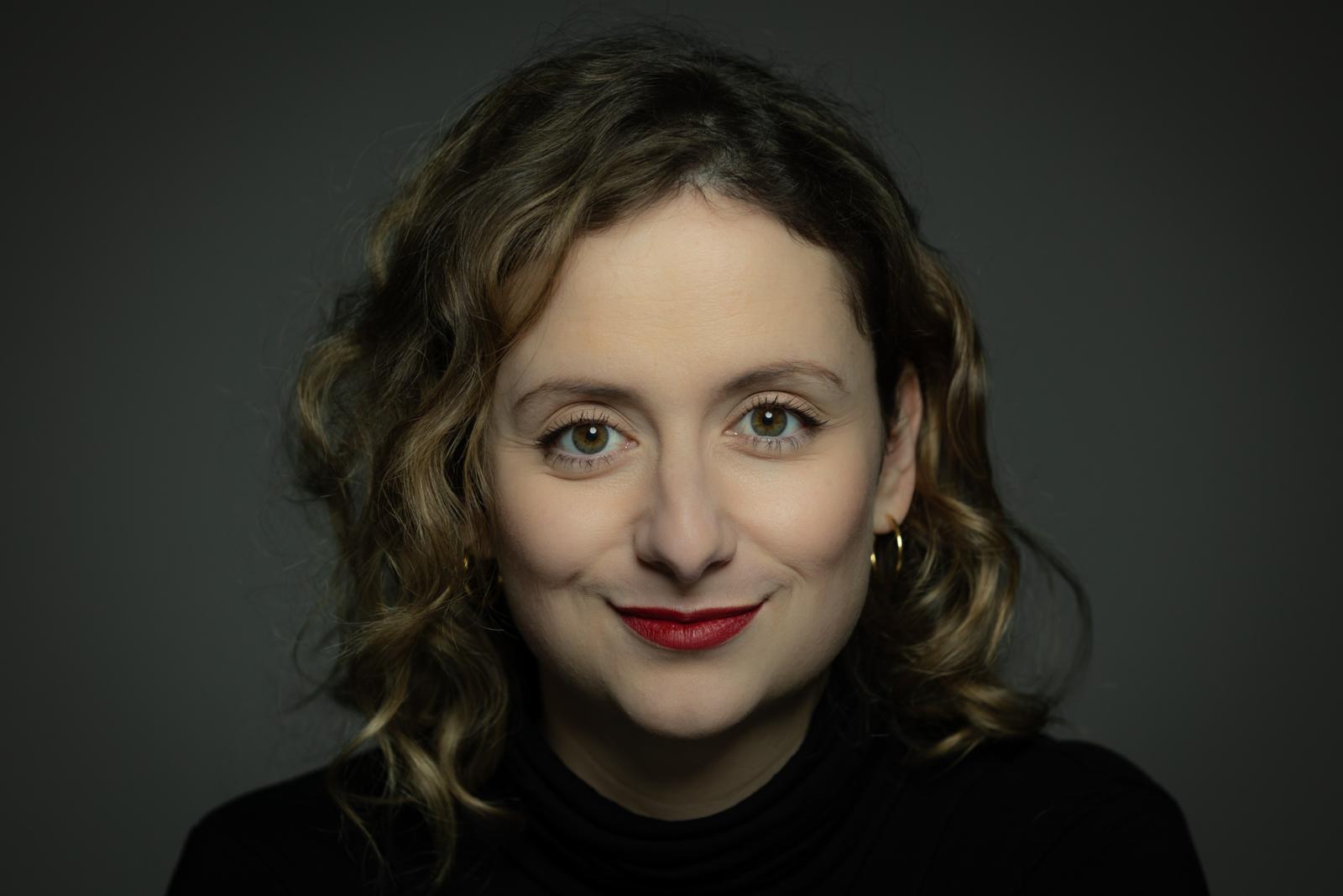 Alexandra Zykonow Nahaufnahme von Alexandra Zykonow mit schulterlangen, lockigen, hellbraunen Haaren. Sie blickt lächelnd in die Kamera, trägt roten Lippenstift, dezente goldene Ohrringe und einen schwarzen Rollkragenpullover vor einem grauen Hintergrund.