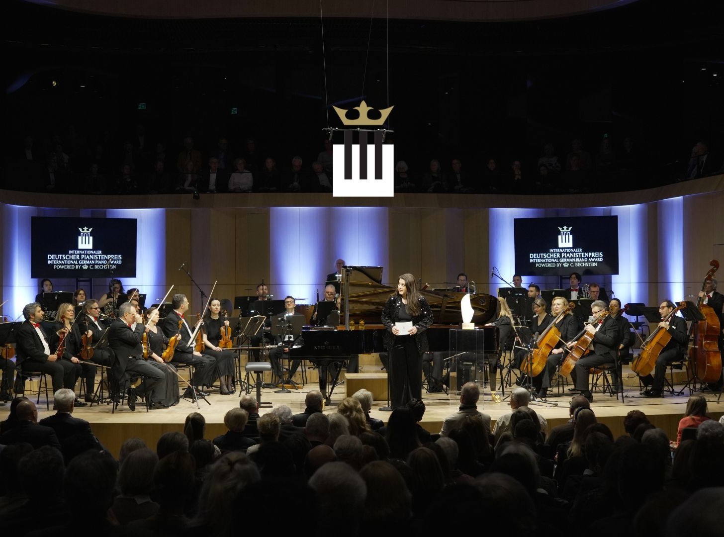 Grand Prix Final Concert Blick auf eine Konzertbühne beim Internationalen Deutschen Pianistenpreis: Eine Sprecherin steht im Scheinwerferlicht im Zentrum, flankiert von einem großen Orchester und einem Konzertflügel, während im Vordergrund das Publikum im Saal sitzt.
