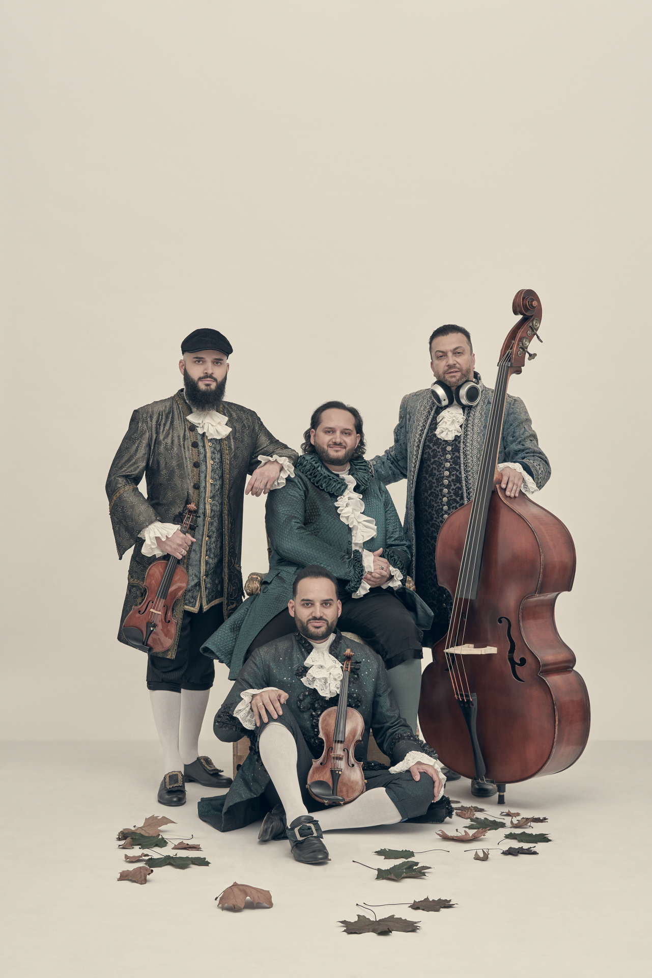 Janoska Ensemble Die vier Musiker des Janoska Ensemble in historisierenden Barock-Kostümen mit Rüschenhemden vor einem hellen, minimalistischen Hintergrund. Zwei Musiker halten Geigen, einer steht neben einem großen Kontrabass und trägt moderne DJ-Kopfhörer um den Hals. Auf dem Boden verstreut liegen vereinzelte Herbstblätter.