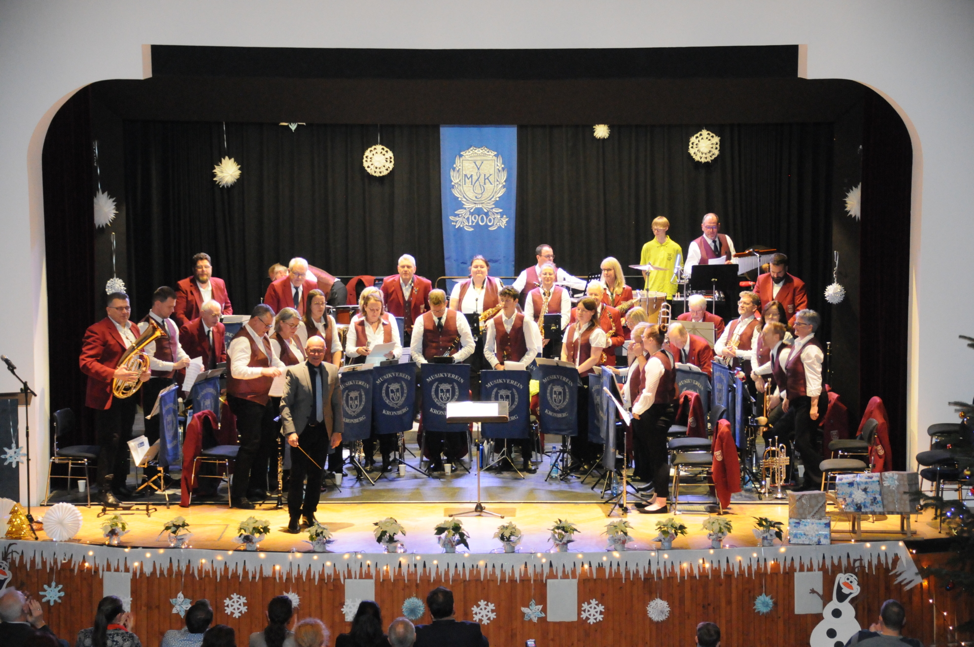 Orchester des Musik-Vereins Kronberg Ein winterlich dekoriertes Konzertfoto zeigt das Orchester des Musik-Vereins Kronberg auf einer hell erleuchteten Konzertbühne. Im Hintergrund hängt ein blaues Banner mit der Aufschrift „MVK 1906“ vor einem schwarzen Vorhang, der mit weißen Sternen verziert ist. Die zahlreichen Musikerinnen und Musiker tragen rote Westen oder Sakkos über weißen Hemden und sitzen hinter dunkelblauen Notenpultbannern mit dem Vereinslogo. Im Vordergrund steht der Dirigent mit erhobenem Taktstock vor dem Orchester. Die Vorderseite der Bühne ist mit einer winterlichen Holzverkleidung, Schneeflocken-Motiven und einer Olaf-Eiskönigin-Figur dekoriert.
