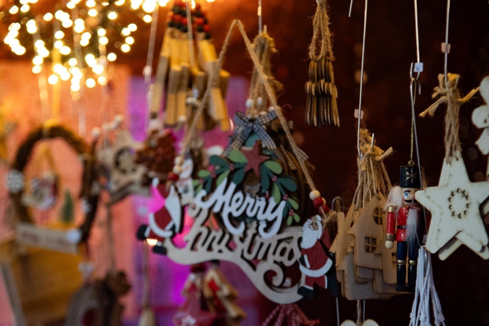 Weihnachtsmarktstand Nahaufnahme von handgemachtem Weihnachtsdekorationen an einem Marktstand, darunter ein Schild mit der Aufschrift „Merry Christmas“, Nussknacker und Holzsterne vor warmen Lichterketten.