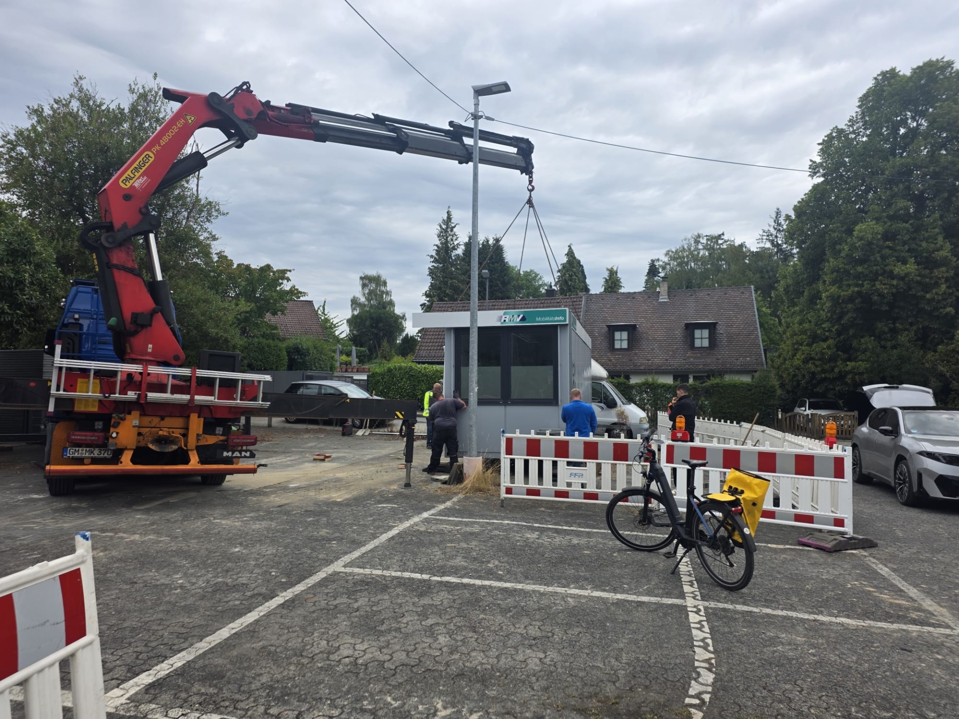 Umzug der RMV-Mobilitätszentrale Ein roter Ladekran eines Lkw hebt ein kleines, graues RMV-Servicehäuschen auf einem Parkplatz an. Bauarbeiter in Arbeitskleidung sichern den Vorgang ab, während im Vordergrund Absperrgitter und ein E-Bike stehen