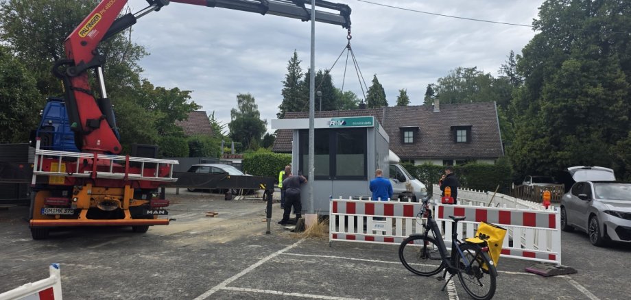 Umzug der RMV-Mobilitätszentrale Ein roter Ladekran eines Lkw hebt ein kleines, graues RMV-Servicehäuschen auf einem Parkplatz an. Bauarbeiter in Arbeitskleidung sichern den Vorgang ab, während im Vordergrund Absperrgitter und ein E-Bike stehen