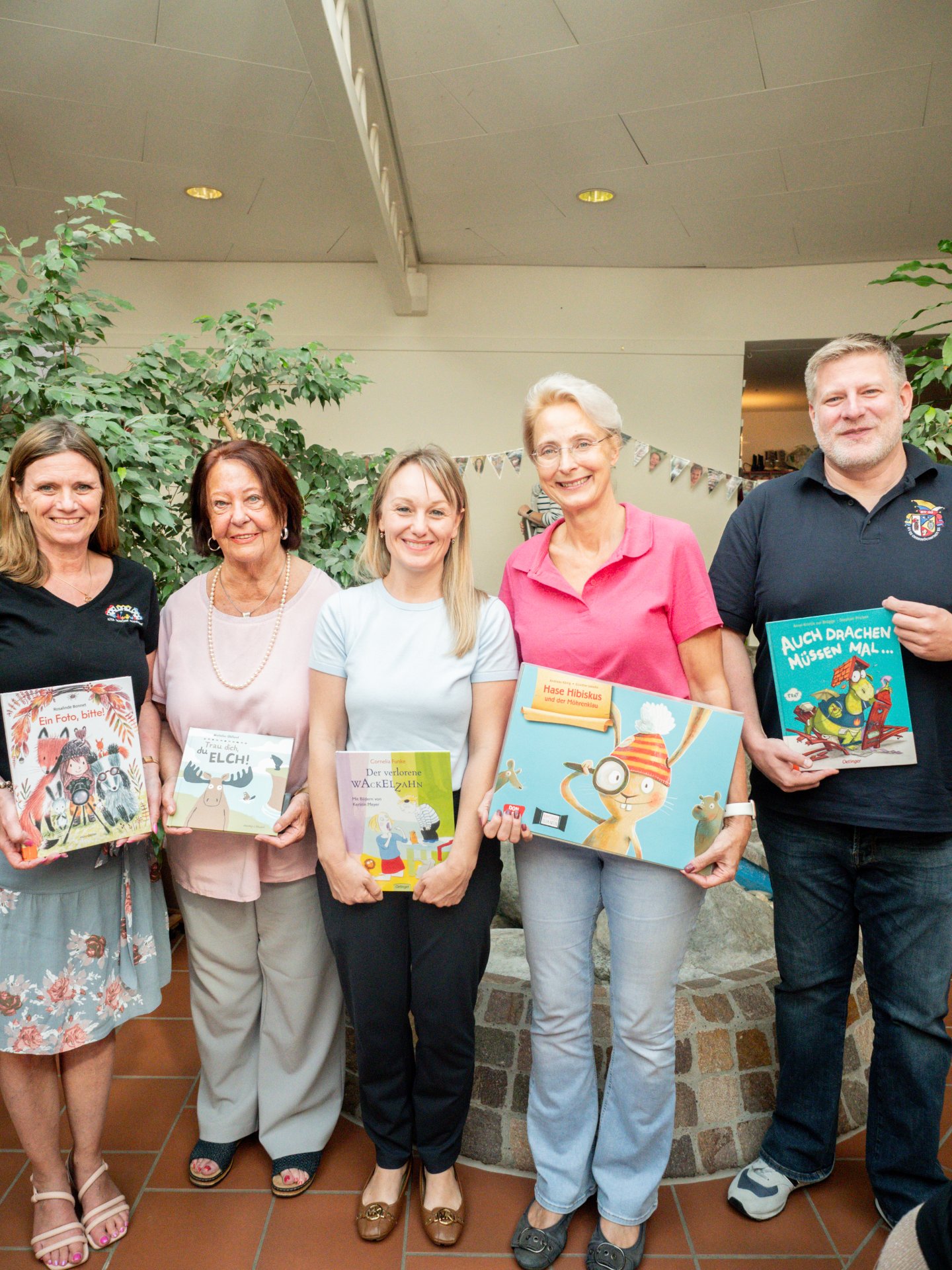 Sie machten mit ihren „Herzensbüchern“ den Vorlese-Nachmittag zu einem besonderen Erlebnis für die Kinder der Kita (v.li.): Gabi Krieger, Doris Bender, Dr. Nicole Pryca, Claudia Opsomer und Jörg Kouth. Ein Gruppenfoto von fünf Erwachsenen (vier Frauen und ein Mann), die nebeneinander in einem Innenraum stehen und lächelnd jeweils ein Kinderbuch in den Händen halten. • Von links nach rechts: • Eine Frau mit dunklen, schulterlangen Haaren in einem schwarzen Oberteil und einem gemusterten Rock hält das Buch „Ein Foto, bitte!“. • Eine ältere Frau mit braunem Bob-Haarschnitt in einem hellrosa Oberteil und grauen Händen hält das Buch „Trau dich, Elch!“. • Eine jüngere Frau mit blonden, halblangen Haaren in einem hellblauen T-Shirt und dunklen Händen präsentiert das Buch „Der verlorene Wackelzahn“. • Eine Frau mit kurzen, graumelierten Haaren und Brille in einem kräftig rosa Poloshirt und Jeans zeigt das größte Buch, „Hase Hibiskus und der Möhrenklau“. • Ein Mann mit kurzen, hellen Haaren in einem dunkelblauen Poloshirt hält das Buch „Auch Drachen müssen mal…“. Im Hintergrund ist ein heller Raum mit Fliesenboden, einer großen Zimmerpflanze auf der linken Seite und einer angedeuteten Steineinfassung (möglicherweise ein Brunnen oder Beet) im unteren Bereich zu sehen