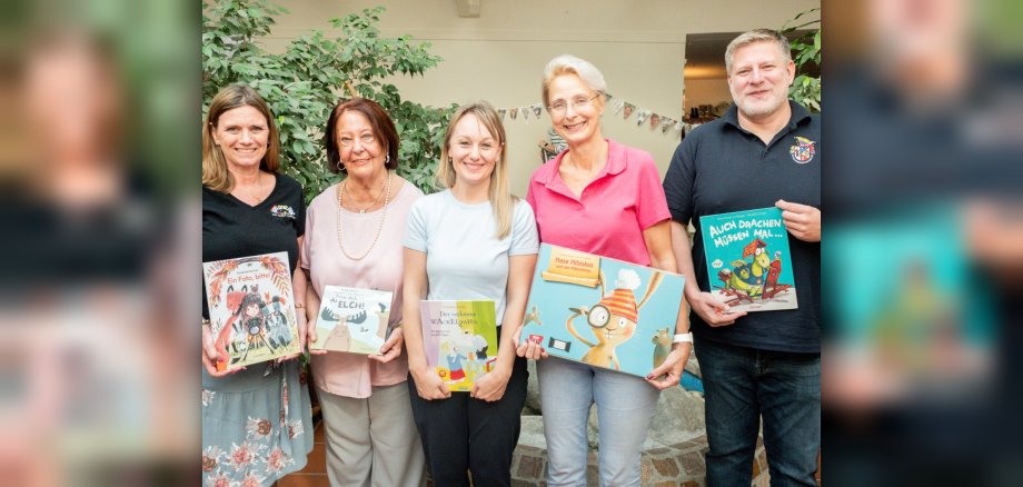 Sie machten mit ihren „Herzensbüchern“ den Vorlese-Nachmittag zu einem besonderen Erlebnis für die Kinder der Kita (v.li.): Gabi Krieger, Doris Bender, Dr. Nicole Pryca, Claudia Opsomer und Jörg Kouth. Ein Gruppenfoto von fünf Erwachsenen (vier Frauen und ein Mann), die nebeneinander in einem Innenraum stehen und lächelnd jeweils ein Kinderbuch in den Händen halten. • Von links nach rechts: • Eine Frau mit dunklen, schulterlangen Haaren in einem schwarzen Oberteil und einem gemusterten Rock hält das Buch „Ein Foto, bitte!“. • Eine ältere Frau mit braunem Bob-Haarschnitt in einem hellrosa Oberteil und grauen Händen hält das Buch „Trau dich, Elch!“. • Eine jüngere Frau mit blonden, halblangen Haaren in einem hellblauen T-Shirt und dunklen Händen präsentiert das Buch „Der verlorene Wackelzahn“. • Eine Frau mit kurzen, graumelierten Haaren und Brille in einem kräftig rosa Poloshirt und Jeans zeigt das größte Buch, „Hase Hibiskus und der Möhrenklau“. • Ein Mann mit kurzen, hellen Haaren in einem dunkelblauen Poloshirt hält das Buch „Auch Drachen müssen mal…“. Im Hintergrund ist ein heller Raum mit Fliesenboden, einer großen Zimmerpflanze auf der linken Seite und einer angedeuteten Steineinfassung (möglicherweise ein Brunnen oder Beet) im unteren Bereich zu sehen