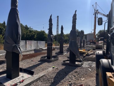 Fundamentstützen Eine Außenbaustelle an Bahngleisen bei sonnigem Wetter. Im Vordergrund stehen mehrere eckige, schwarze Fundamentstützen, deren obere Hälften mit grauer Schutzfolie eingewickelt sind. Rechts im Hintergrund sind ein gelber Baukran und ein Teil eines Baustellenfahrzeugs zu sehen
