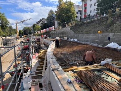 Betonarbeiten an der Stützwand Eine langgezogene Baustelle an einem Hang mit Wohngebäuden im Hintergrund. Mehrere Bauarbeiter arbeiten an der Bewehrung für ein Betonfundament, indem sie Stahlmatten verlegen. Links befinden sich Baugerüste, ein kleiner roter Bagger und im Hintergrund ein gelber Kran unter leicht bewölktem Himmel